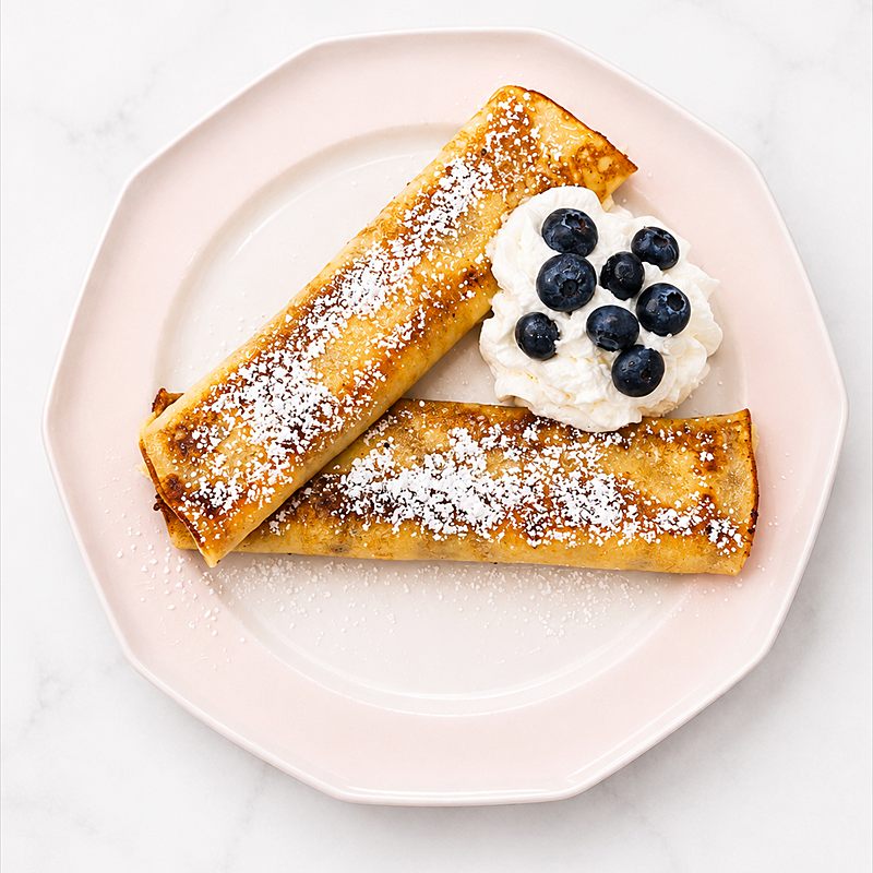 Two rolled blintzes dusted with icing sugar, with cream and blueberries.